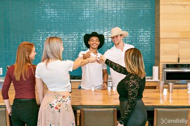 Five friends toasting with small shot cups across a wooden kitchen island in front of a teal tile backsplash, two men wearing cowboy hats and everyone smiling.