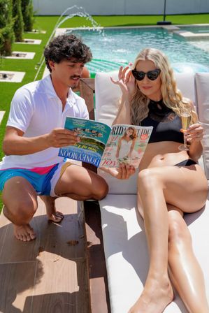 Two adults relaxing poolside on a sunny day: a woman in a black bikini and cat-eye sunglasses sips champagne and reads a wedding magazine while a man in a white polo and colorful swim trunks crouches beside her, resort-style backyard pool and fountain in background.