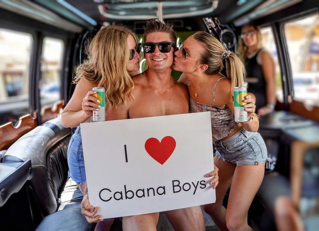 Three friends in a party van: a shirtless man in sunglasses holds a sign with a red heart supporting a cabana crew while two women kiss his cheeks and hold canned drinks.