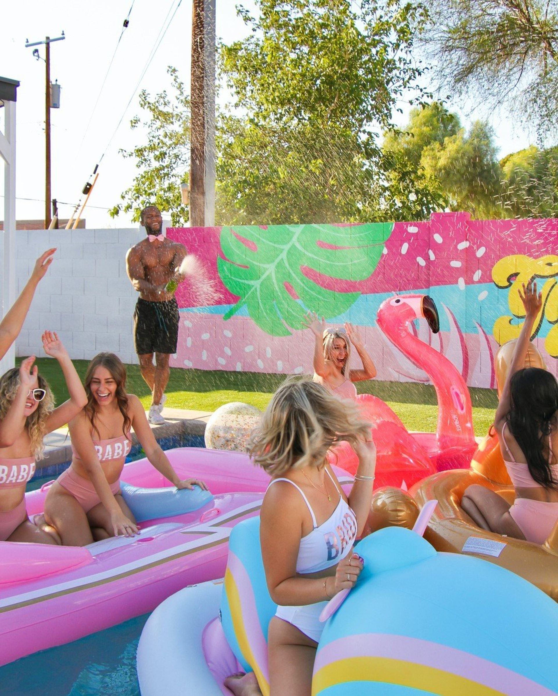 Sunlit backyard summer pool party with friends on colorful inflatable floats—pink flamingo and pastel rides—laughing women in swimsuits as a shirtless man sprays water by a tropical mural
