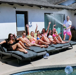 Sunny backyard pool party with a row of women in swimsuits cheering on poolside loungers as a man sprays champagne over them, water droplets sparkling in the air.