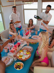 Friends in swimsuits clinking champagne flutes at an indoor summer brunch around a turquoise table with pink candles, floral centerpieces, emoji cookies, fruit bowl and servers bringing fried chicken.