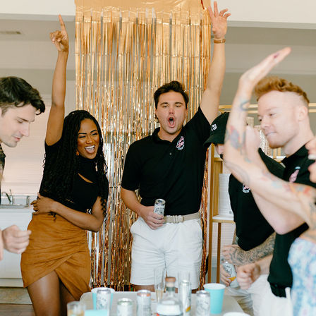 Friends cheering with hands raised at an indoor party, gold fringe backdrop and canned drinks on the table.