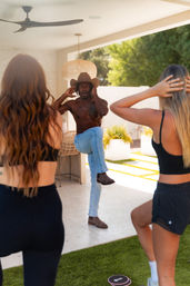 Shirtless instructor wearing a cowboy hat leads an outdoor balance workout on a sunny modern backyard patio while two women in activewear mirror a one-leg pose