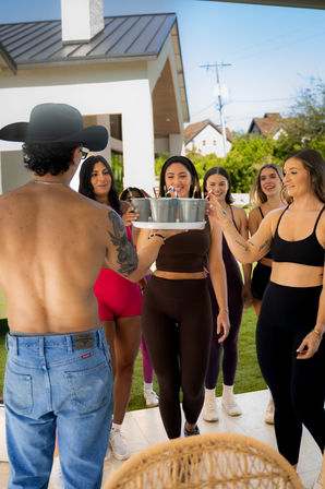 Smiling group of women in colorful workout clothes on a sunny suburban backyard patio taking drinks from a shirtless man wearing a cowboy hat.