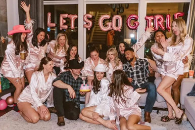 Lively bachelorette party in a cozy living room with women in matching pink satin robes and two men, metallic balloon letters reading LET S GO GIRLS, cowboy hats, a small cake and festive celebration vibes