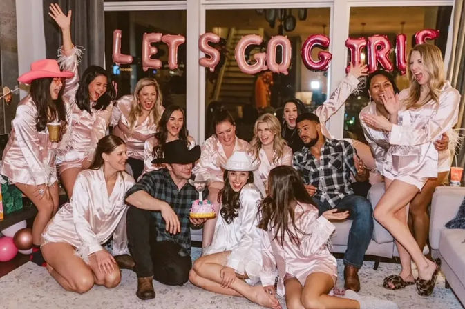 Lively bachelorette party in a cozy living room with women in matching pink satin robes and two men, metallic balloon letters reading LET S GO GIRLS, cowboy hats, a small cake and festive celebration vibes