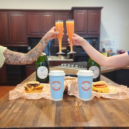 Two people toasting mimosas over a wooden kitchen island with takeaway coffee cups and breakfast sandwiches — casual at-home brunch scene.
