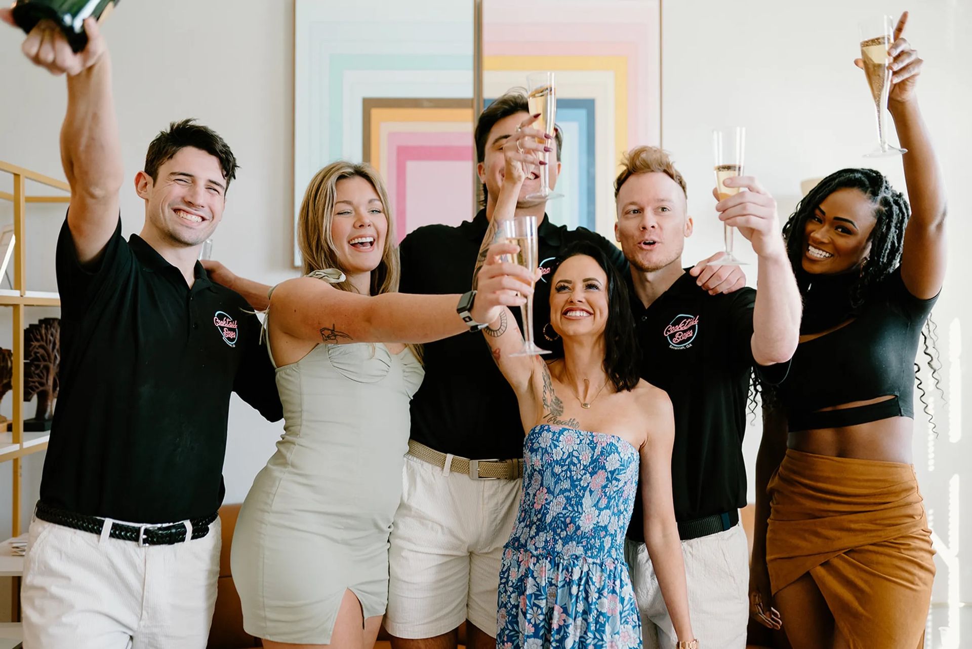 Six friends in casual summer outfits raise champagne flutes for a cheerful indoor toast in a modern living room with colorful geometric wall art.