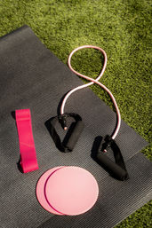 Outdoor fitness gear on a black yoga mat: pink loop resistance band, pink resistance tube with black foam handles, and two pink gliding discs on green grass