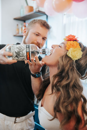 Man pouring liquor from a bottle into a woman’s open mouth while she wears a colorful flower crown at an indoor party with balloons
