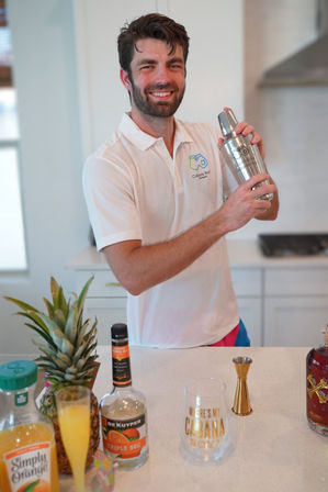 Smiling man in a white polo shaking a cocktail shaker at a bright modern kitchen counter with pineapple, orange juice, liqueur bottles and a jigger.