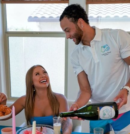 Smiling woman in a blue dress at a sunlit dining table as a server in a white polo pours sparkling wine for a casual brunch celebration with appetizers and candles.