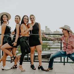Playful rooftop party: four women in chic black outfits pose with champagne while a cowboy-hatted man playfully lassos them against an urban city skyline.