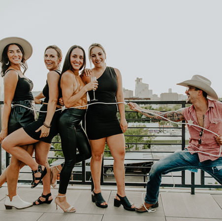Playful rooftop party: four women in chic black outfits pose with champagne while a cowboy-hatted man playfully lassos them against an urban city skyline.