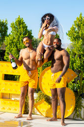 Sunny poolside bachelorette scene: bride-to-be in white bikini and veil with heart sunglasses lifted by two shirtless friends in orange swim trunks, one holding champagne, yellow marquee letters and greenery in background.
