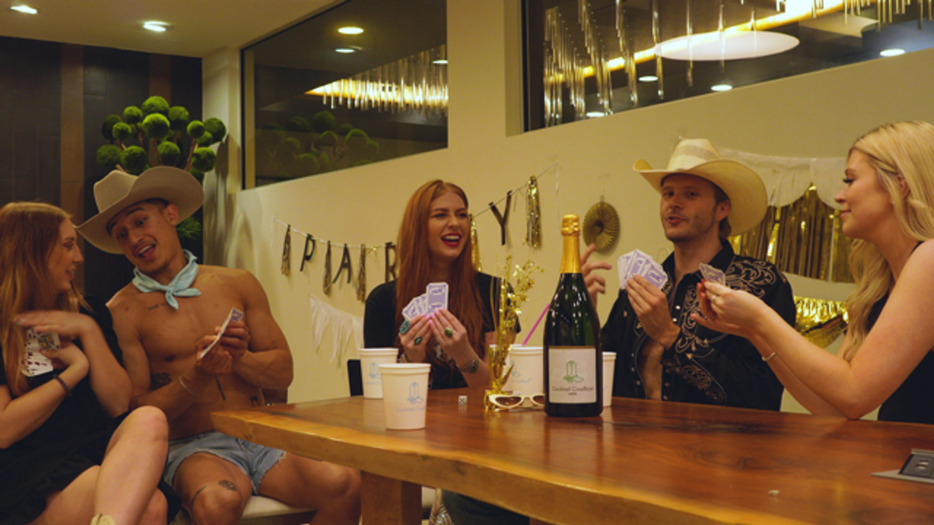 Group of young adults at an indoor living-room party playing cards around a wooden table, champagne bottle and cups on the table, gold streamers and a festive banner in the background, some wearing cowboy hats.