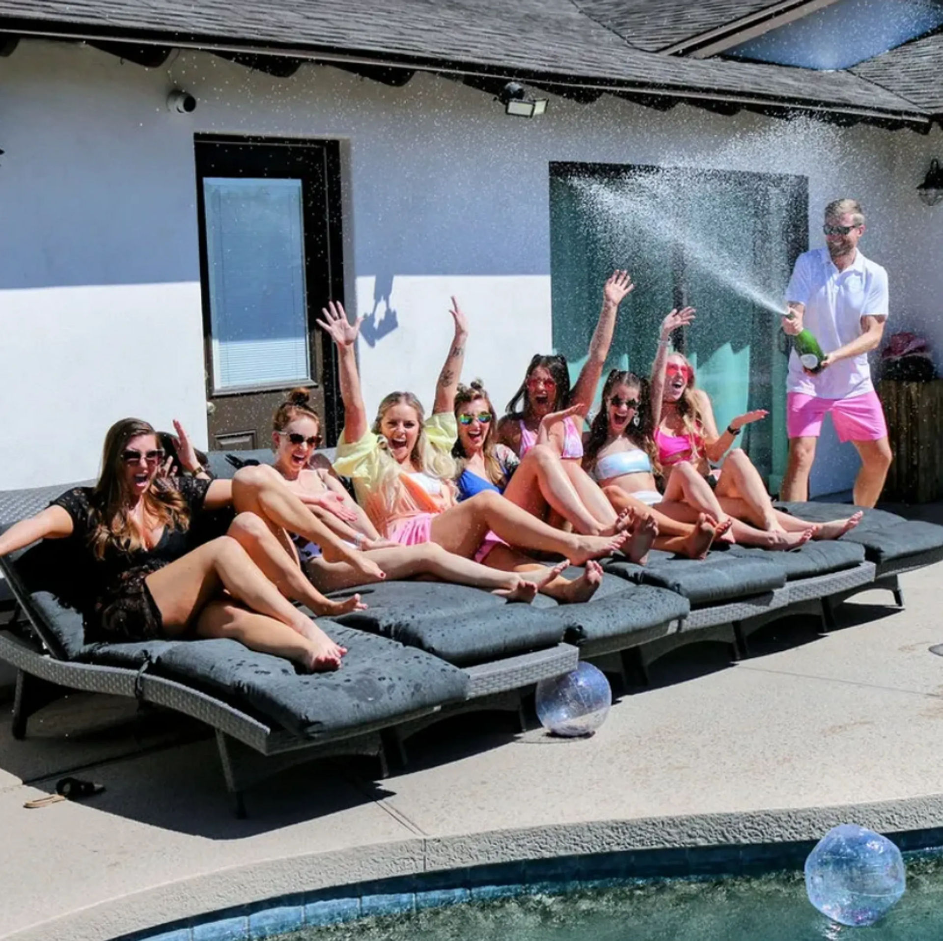 Sunlit backyard pool scene: eight women in colorful swimsuits cheering on cushioned lounge chairs while a man sprays champagne, splashing across the pool deck.