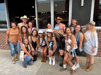 Cheerful group of women and two shirtless men in cowboy hats posing on a city sidewalk outside a brick storefront, many wearing cowboy boots, bandanas and festive outfits — playful country-night out photo.