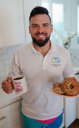 Smiling man in a bright kitchen holding a coffee mug that reads "YOU'RE LIKE, REALLY PRETTY" and a plate of seeded bagels — casual breakfast scene.