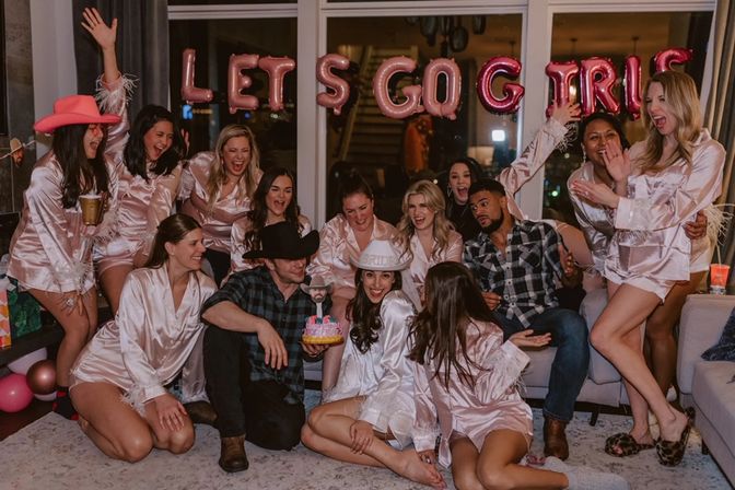 Bachelorette party in a living room with friends in pink satin robes and cowboy hats laughing and cheering around the bride holding a small cake under a balloon banner reading 'LET'S GO GIRLS'.