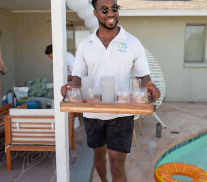 Smiling man in sunglasses carrying a wooden tray of glasses poolside at a sunny backyard party, with an inflatable orange ring and patio seating in the background.