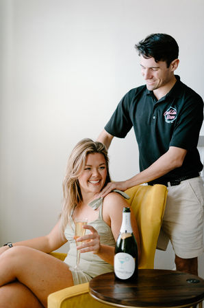 Smiling woman in a light dress sits in a yellow armchair holding a champagne flute while a man in a black polo gives a shoulder massage, champagne bottle on a side table—relaxed indoor celebration scene.
