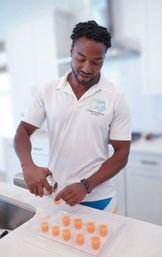 Man pouring orange cocktail shots from a shaker into a tray of plastic shot cups on a white modern kitchen island, bright home bar setting.