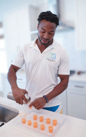 Person in white polo pouring orange cocktail shots from a shaker into small plastic cups on a clear tray on a bright modern kitchen countertop
