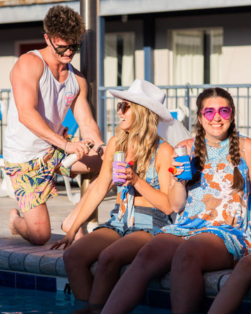 Sunny poolside party with friends: a man in tropical shorts opens a drink while two women sit on the pool edge with canned drinks—one wearing a white cowboy hat and sunglasses, the other in heart-shaped pink shades and a blue floral dress.