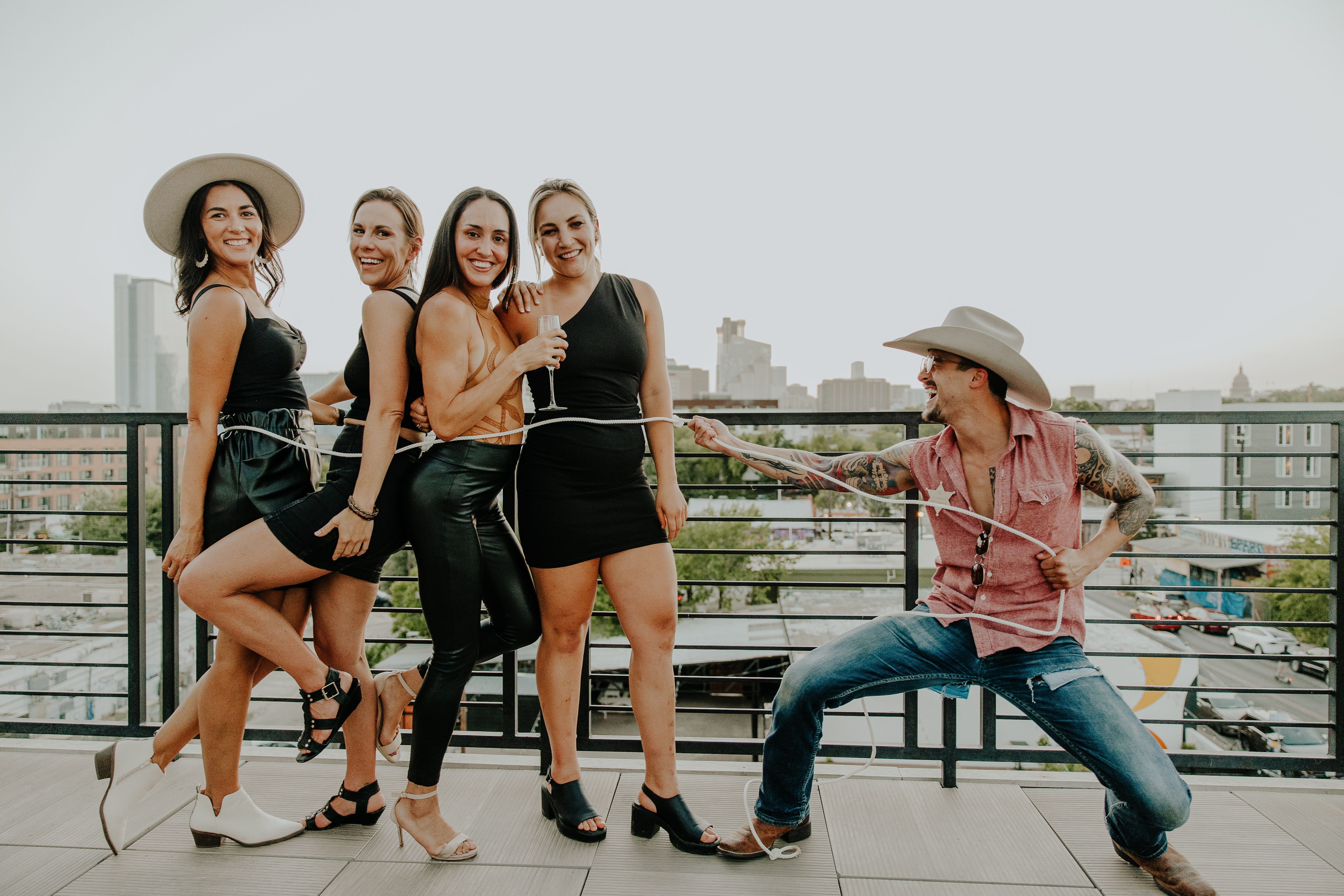 Four women in black outfits pose on an urban rooftop terrace, smiling and holding a champagne glass while a man in a cowboy hat playfully lassos them with a rope against a city skyline.