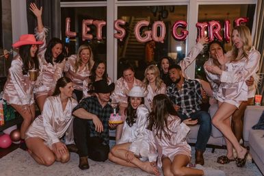 Bachelorette party in a living room: bride-to-be in white satin robe and cowboy hat surrounded by friends in matching pink satin robes, holding a small cake under pink “LET'S GO GIRLS” balloons, everyone cheering.