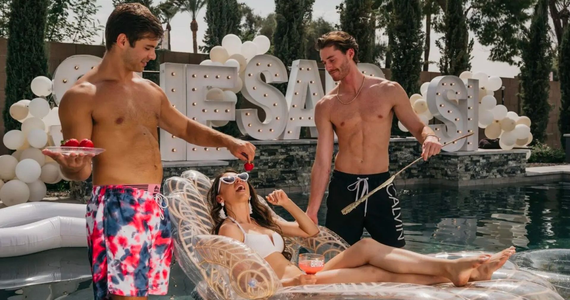 Two shirtless men feed strawberries to a woman lounging on a clear inflatable at a sunny backyard pool party with white balloons and lit marquee letters — playful bachelorette vibe.