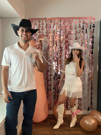 Indoor party photo of a man in a black cowboy hat and white polo playfully holding a lasso tied around a surprised woman in a white cowgirl fringe dress, white hat and boots, posing in front of a rose-gold metallic fringe backdrop with balloons.
