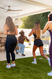 Backyard outdoor fitness class on a covered patio: shirtless instructor wearing a cowboy hat and jeans leading three women in athletic wear through lunges on artificial turf, with a ceiling fan, woven pendant light and a grill in the background.