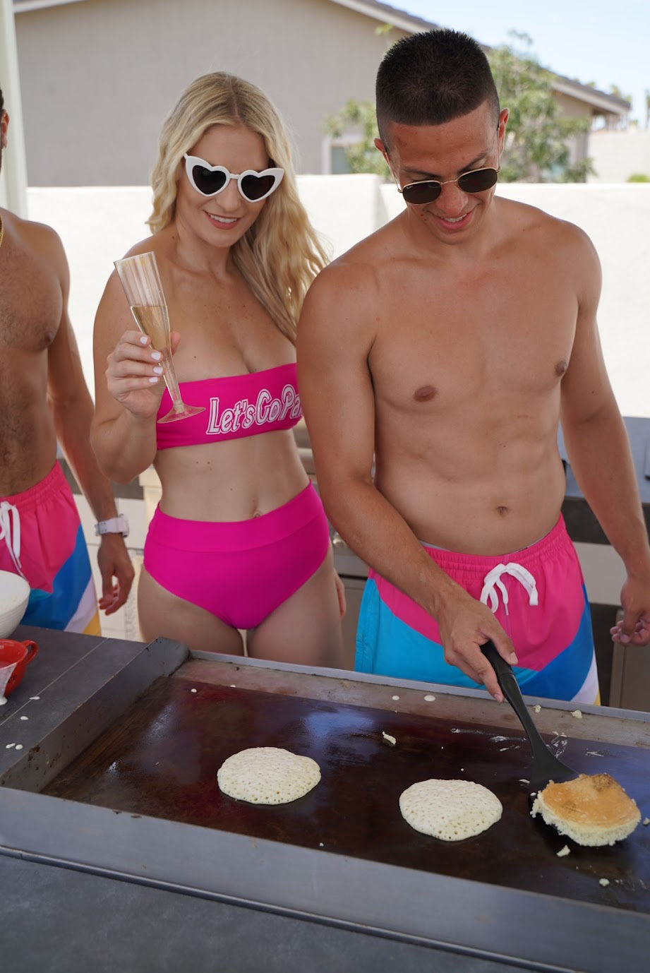 Two adults in bright pink swimwear cooking pancakes on a backyard griddle at a sunny poolside summer brunch — woman in heart-shaped sunglasses holds a champagne flute while a shirtless man flips pancakes.