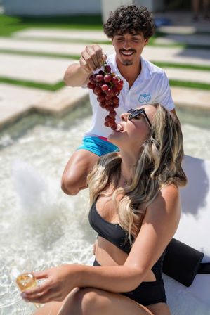 Poolside scene: smiling man holds a bunch of red grapes above a woman in a black swimsuit and sunglasses as she leans back to bite one, lounging on a sunlit pool ledge with a drink in hand.