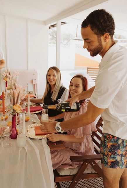 Sunlit covered patio brunch with a man pouring sparkling wine for two smiling women at a pastel floral tablescape with candles and place settings