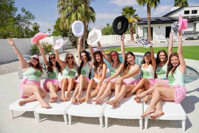 Poolside bachelorette party with a row of women in matching mint tank tops and pink skirts cheering and waving cowboy hats by a sunny backyard pool with palm trees.