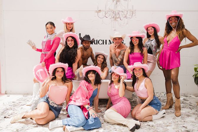 Group of partygoers in pink cowgirl outfits posing around a bathtub prop in a bright white studio, wearing fluffy pink cowboy hats and boots amid scattered play money under a chandelier.