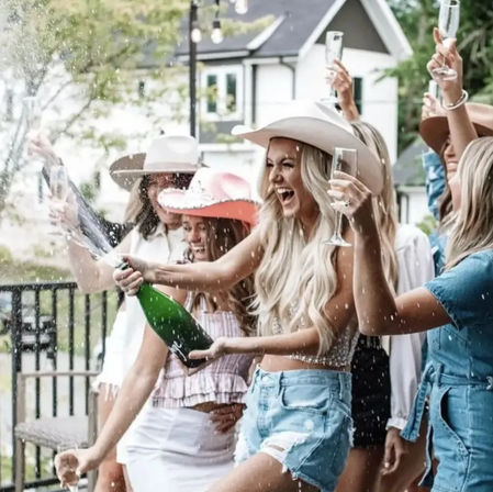 Group of women at a suburban backyard bachelorette party popping champagne and cheering in cowboy hats on a sunny patio.
