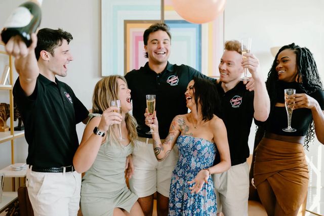 Six friends laughing and toasting with champagne flutes at an indoor celebration, wearing casual summer outfits with colorful geometric wall art and a balloon in the background.