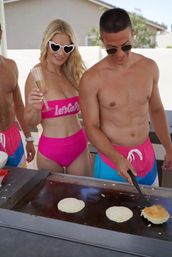 Sunny backyard poolside brunch: three friends in colorful swimwear cooking pancakes on a flat-top griddle — woman in pink high-waisted bikini and heart sunglasses holds a champagne flute while a shirtless man flips a pancake.