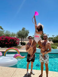 Sunny backyard pool party: two shirtless men lift a woman in a white "wifey" swimsuit and sunhat as she raises a pink funnel drinking tube, with a rainbow inflatable in the turquoise pool and palm trees in the background.