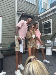 Three friends laughing on a wooden deck beside painted clapboard houses — two in pink polos and white sneakers and a woman in a crochet beach outfit holding a cocktail, casual summer backyard party vibe.