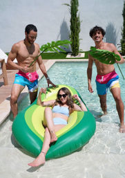 Three friends at a sunny backyard pool — woman in light blue bikini lounging on an inflatable avocado float with a drink while two shirtless men in colorful swim trunks playfully hold large tropical leaves, summer pool party vibe.