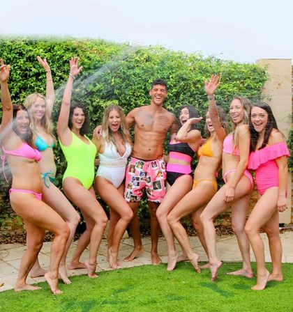 Group of friends in bright swimwear laughing and posing under a sprinkler in a sunny backyard garden, vibrant summer pool-party vibe.