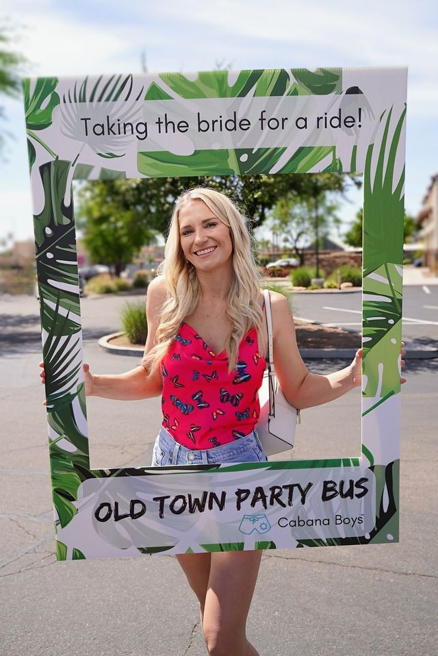 Smiling woman in a pink butterfly top holding a tropical photo-frame prop that reads "Taking the bride for a ride!" in a sunny suburban parking lot at a bachelorette-party celebration.