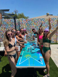 Group of friends in bikinis cheering with cups around a blue beer-pong table at a sunny backyard summer party, wearing heart-shaped sunglasses in front of a colorful heart-pattern mural.