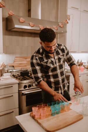 Man pouring clear liquor into colorful plastic shot glasses on a wooden board in a modern home kitchen with stainless steel stove and festive garland